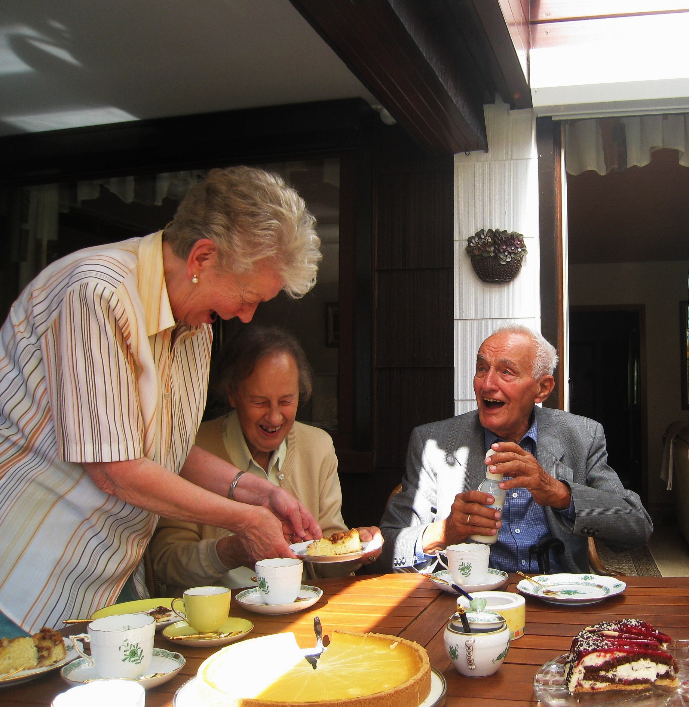 Das Bild zeigt eine Seniorin, die zwei Senioren ein Stück Kuchen reicht.